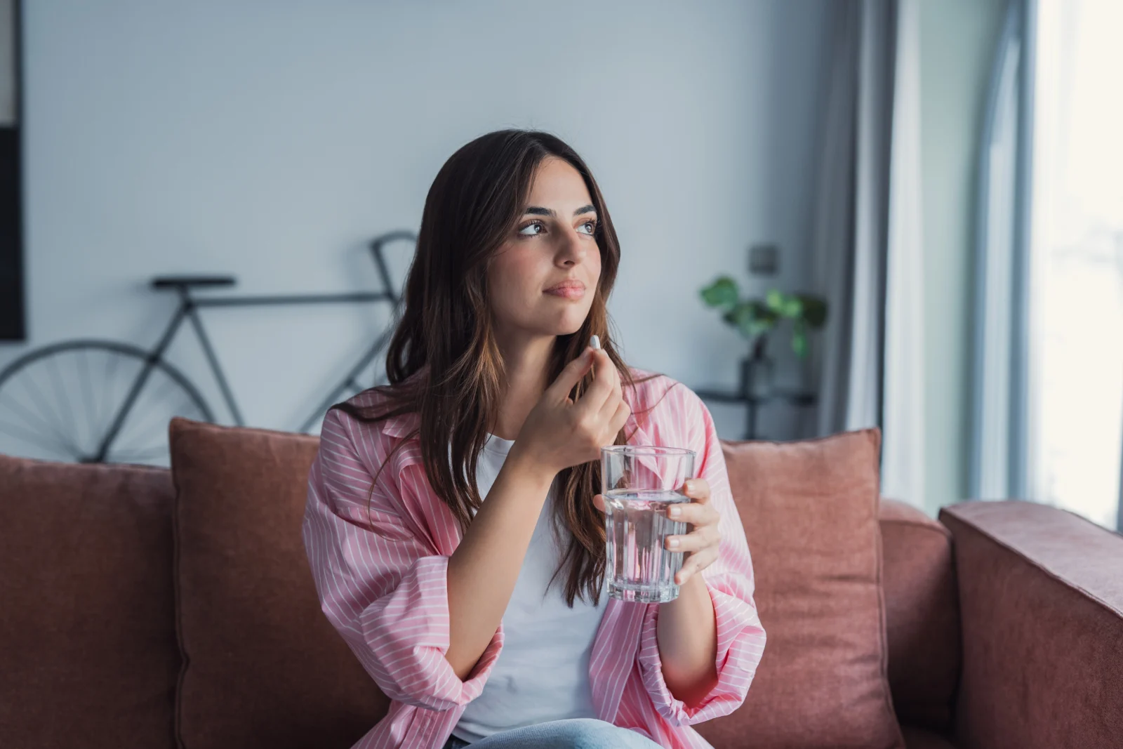 Woman sitting on a couch holding a glass of water, appearing thoughtful while considering medication options